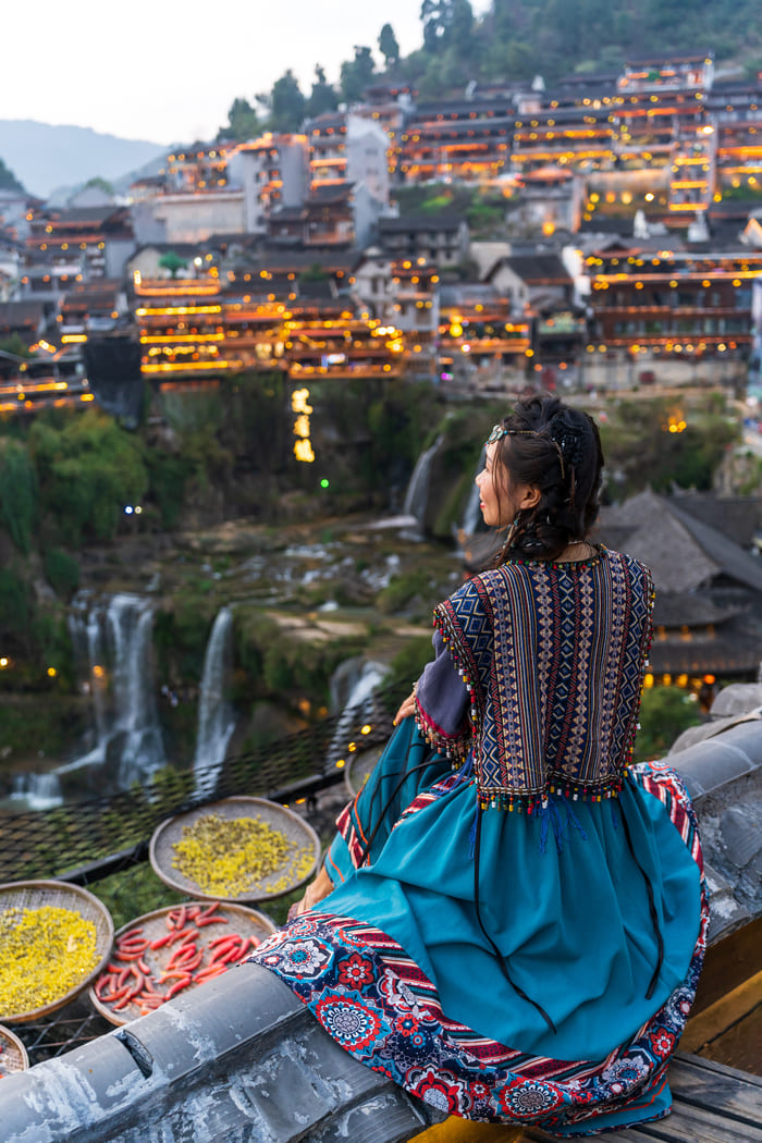 Mujer de espalda vistiendo un traje tradicional colorido y falda azul turquesa, contemplando un pueblo iluminado en la montaña